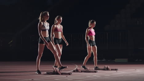 Eine-Reihe-Läuferinnen-Hockt-In-Der-Startposition,-Bevor-Sie-Mit-Dem-Rennen-Beginnen.-Frauen-Starten-Mit-Laufschuhen-Im-Stadion-Von-Der-Startlinie-Im-Dunkeln-Mit-Scheinwerfern-In-Zeitlupe.